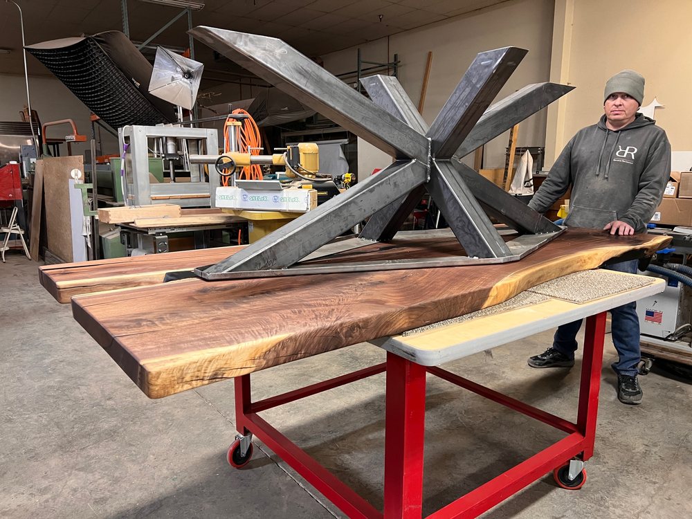 Multi-arm steel table base standing beside a live edge top in the shop, a person alongside for scale