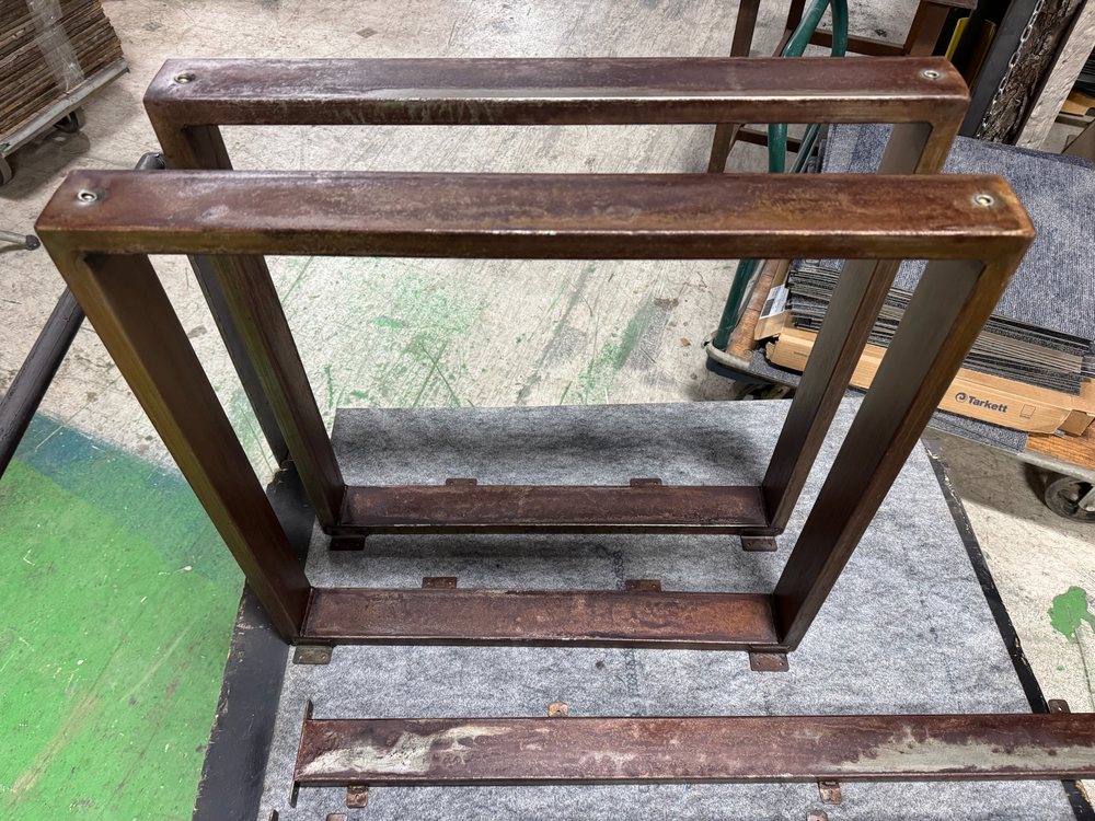 Two rectangular steel base frames leaning together, warm force patina finish under the seal